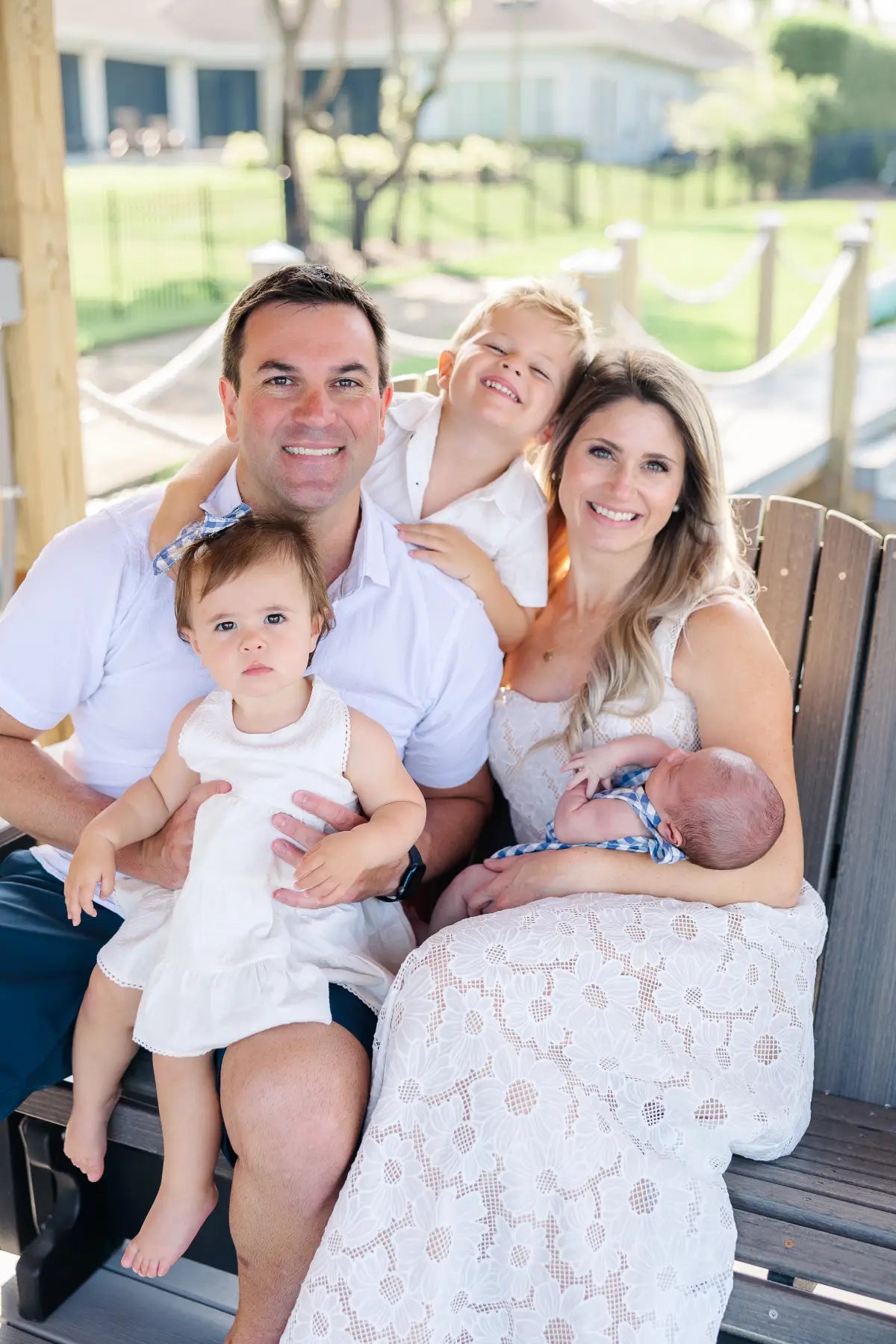 Family of four outdoors on a wooden bench with a house and greenery in the background