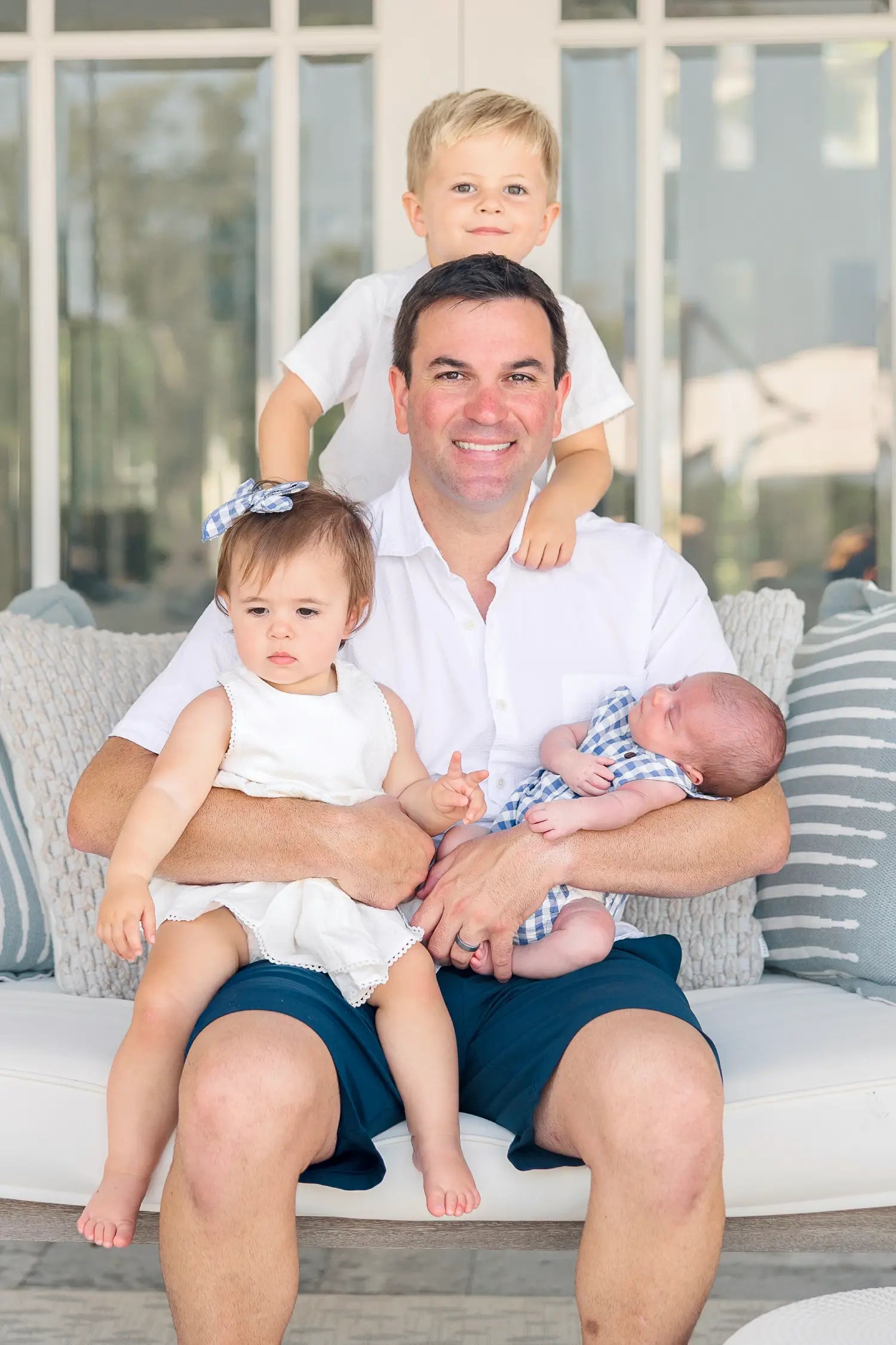 Man sitting on a couch with three children, including two toddlers and a baby, in a bright outdoor setting